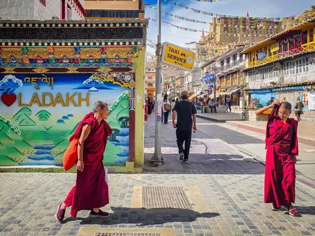 Vibrant Leh Market street scene with monks in maroon robes, colourful “I Love Ladakh” wall art, prayer flags and bustling shops under clear blue sky, ideal for Ladakh culture and street photography blogs