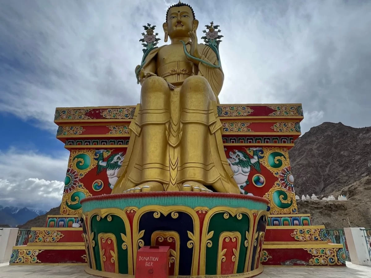 Likir Gompa Ladakh towering 75ft golden Maitreya Buddha statue seated meditative pose coiled naga serpent arm ornate multicolored pedestal shrine red donation box snowy barren Ladakh mountain backdrop cloudy sky monastery buildings, iconic Himalayan spiri
