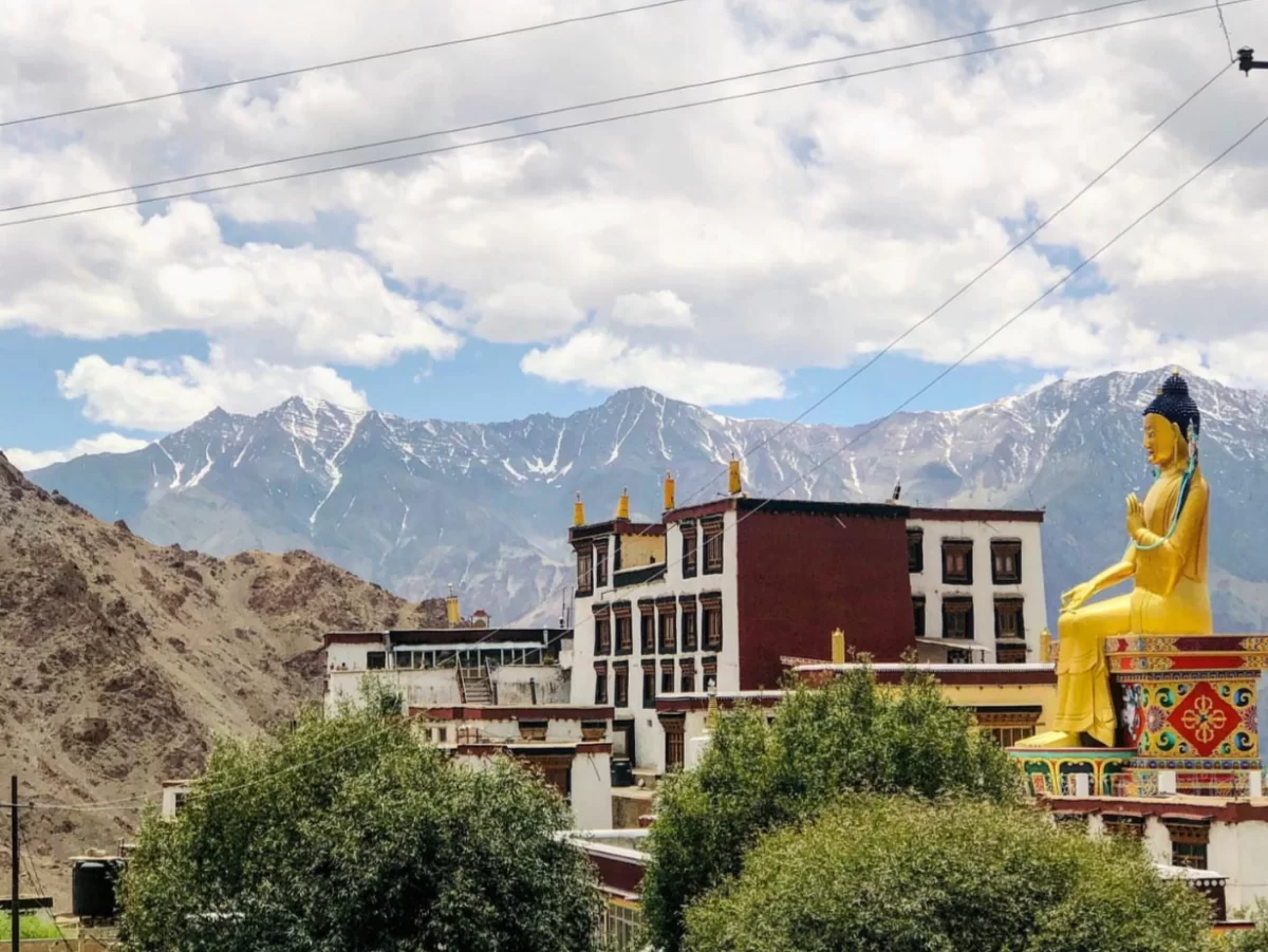 Likir Gompa Ladakh panoramic view golden seated Maitreya Buddha statue atop Gelugpa monastery complex red white buildings snowy Himalayan mountains barren valley trees blue cloudy sky, iconic Buddhist monastery landmark tour package.