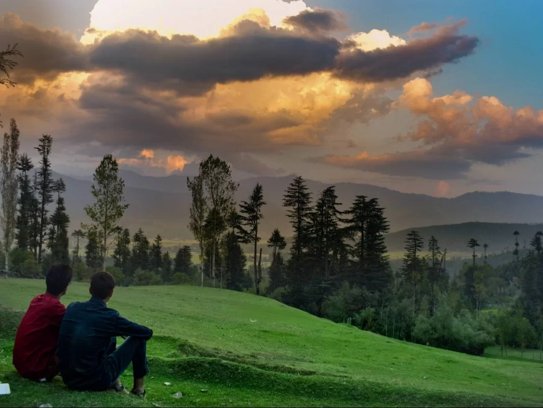 Friends relaxing at Lolab Bungus Valley during dramatic sunset clouds, featuring pine trees distant mountains green meadows, perfect adventure experience Kashmir tour packages.