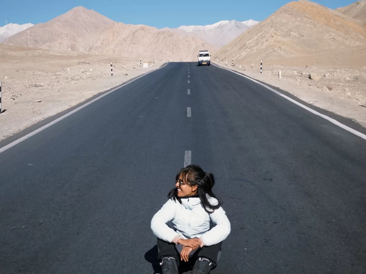 Magnetic Hill optical illusion in Ladakh where white-clad biker sits on roadside overlooking straight asphalt highway flanked by barren brown hills and distant snow mountains with white van ahead, perfect India adventure tour package. 
