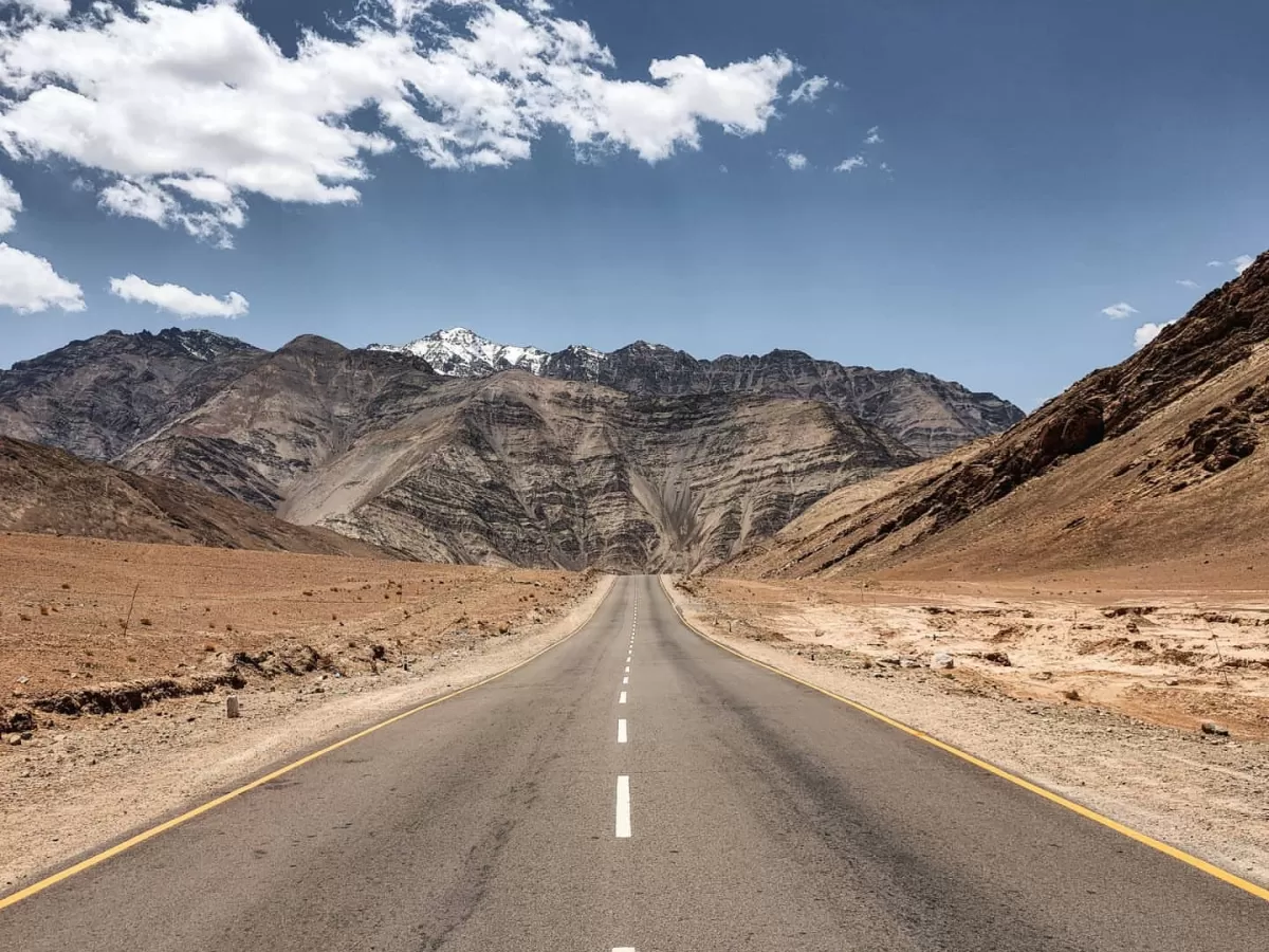 Magnetic Hill Leh Ladakh straight asphalt highway vanishing into distant snow-capped rugged mountains under clear blue skies with scattered white clouds and barren desert landscape, gravity-defying optical illusion road perfect India adventure tour packag
