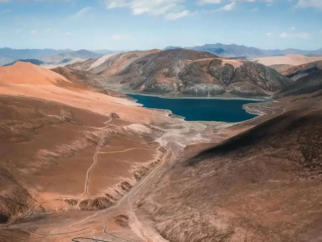 Stunning aerial drone view of turquoise Mirpal Tso Lake nestled amid barren rusty brown desert sand dunes rugged arid mountains in remote high-altitude Changthang Ladakh under clear blue skies, perfect India Leh Ladakh offbeat adventure tour package. 