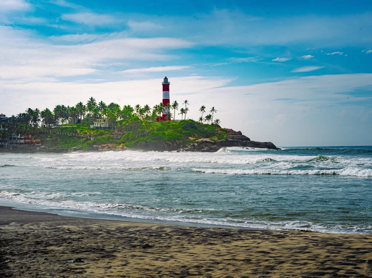 Vibrant blue sky view of Kovalam Beach Kerala featuring red-white lighthouse atop palm-covered rocky cliff, crashing Arabian Sea waves on dark sandy shore, perfect India tour package.