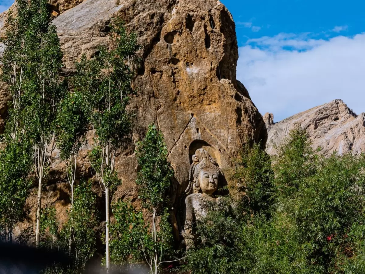 Colossal ancient rock-carved Maitreya Buddha statue emerging from rusty cliff face surrounded by lush green poplar trees against dramatic barren Ladakh mountains blue skies at Mulbekh Monastery near Kargil, perfect India Leh Ladakh Buddhist heritage tour 