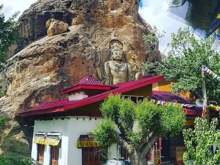 Towering 9-meter rock-carved Maitreya Buddha Future Buddha statue atop Mulbekh Gompa monastery with red-tiled roof white walls fluttering prayer flags lush green trees rugged brown cliffs cloudy skies Ladakh India, perfect Leh Kargil Himalayas Buddhist ro