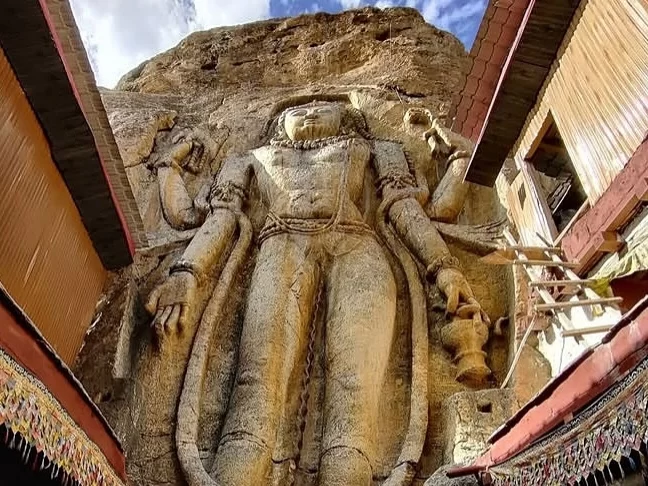 Massive rock-carved 8-meter Avalokiteshvara Padmapani Bodhisattva statue framed by wooden balconies prayer flags Mulbekh Gompa monastery rugged cliffs blue skies Ladakh, perfect Kargil Leh Himalayas Buddhist rock art tour. 