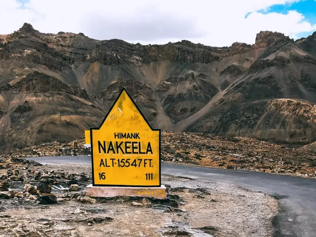 Nakeela pass Himank signboard Ladakh during partly cloudy day, featuring yellow BRO plaque alt 15647 ft, barren rocky mountains winding road, perfect adventure Ladakh tour package.