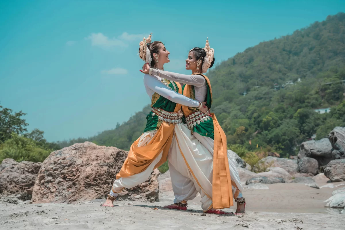 Two dancers in traditional Odissi attire perform a graceful pose on a rocky riverbank against a backdrop of lush green hills and a clear sky. A vibrant cultural display featured in our Odisha packages, celebrating the rich heritage of the region.