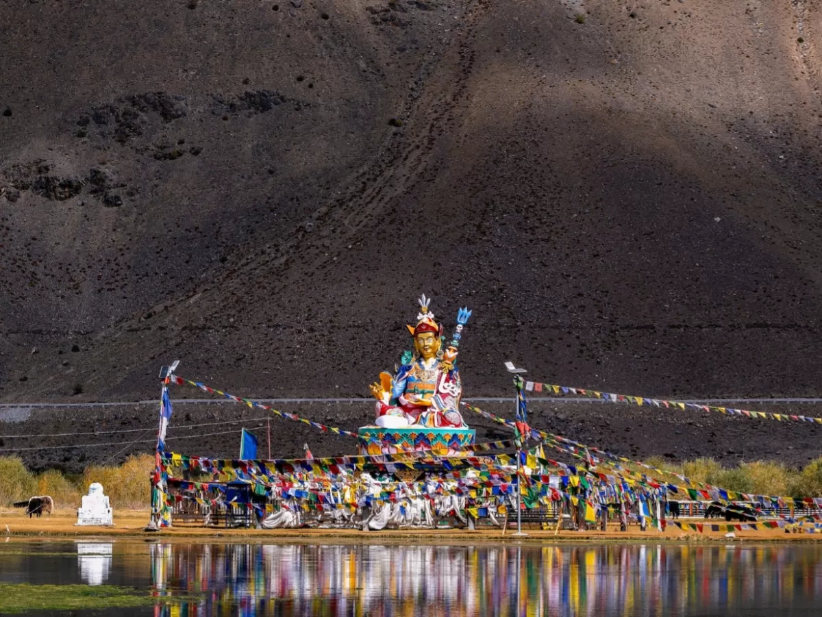 Guru Padmasambhava statue at Sani Lake near Padum village Zanskar Valley Ladakh during clear day, featuring prayer flags, motorcycles, barren mountain backdrop, perfect spiritual Zanskar tour package. 