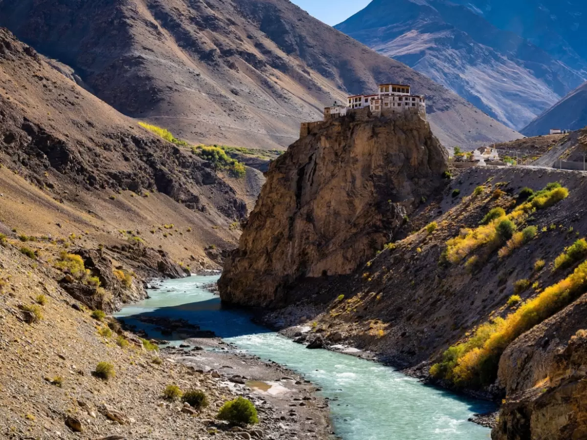 Padum Village Zanskar Ladakh distant white hilltop monastery houses clinging to rugged brown cliffs foreground mani prayer stones painted yellow blue green scattered field, high-altitude Buddhist village landscape tour package.