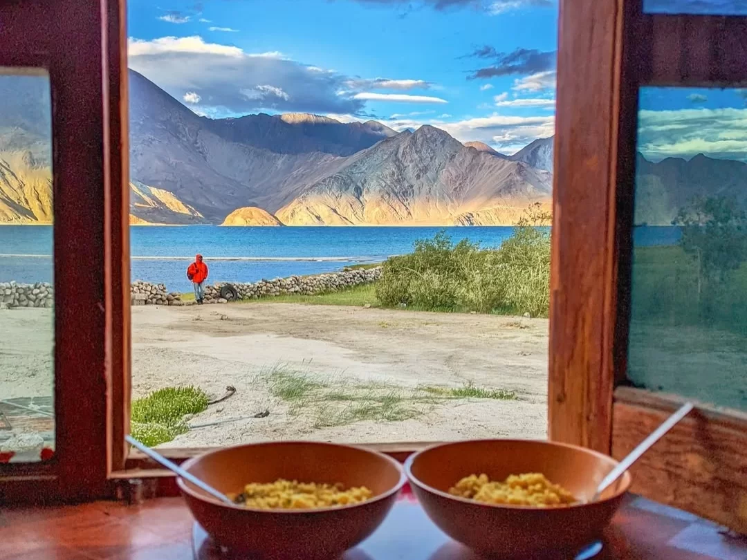 Ladakh Pangong Tso Lake window view turquoise blue brackery water red jacket person on sandy shore snow capped rugged brown Himalayan mountains grassy patch wooden window frame bowls spoons foreground adventure tour package