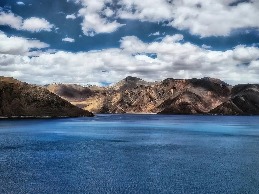 Ladakh Pangong Tso Lake deep blue turquoise brackish water terracotta brown layered rugged Himalayan mountains scattered white clouds clear blue sky high altitude scenic panoramic adventure tour package Ladakh Himalayas. 