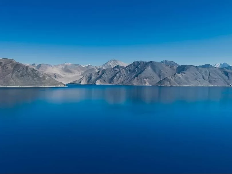 Ladakh Pangong Tso Lake deep blue turquoise brackish water gray rocky snow capped Himalayan mountains clear blue sky high altitude scenic landscape panoramic view adventure trekking tour package Ladakh Himalayas. 