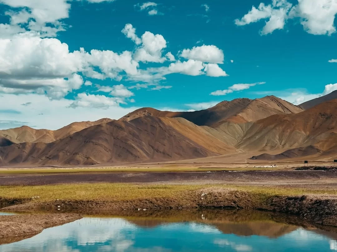 Ladakh Pangong Tso Lake turquoise blue brackish water reflection brown terracotta rugged Himalayan mountains scattered white clouds clear blue sky barren landscape high altitude scenic adventure trekking tour package.