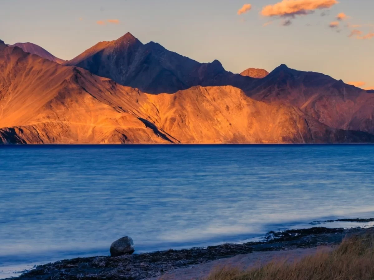 Ladakh Pangong Tso Lake sunset golden orange light rugged snow capped Himalayan mountains turquoise blue brackish water rocky shore grass tufts dramatic high altitude landscape adventure tour package.
