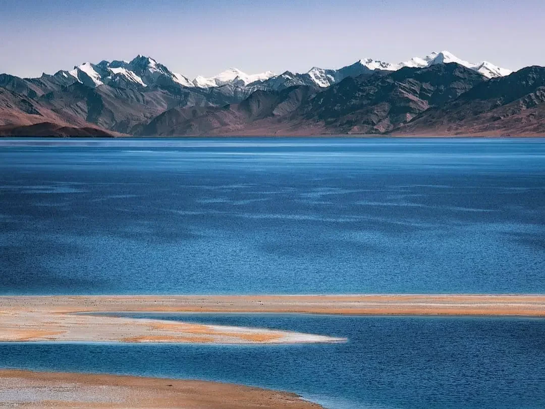 Ladakh Pangong Tso Lake turquoise blue high altitude brackish water snow capped rugged Himalayan mountains brown barren slopes sandy beige shoreline clear sky scenic adventure tour package Ladakh Himalayas. 