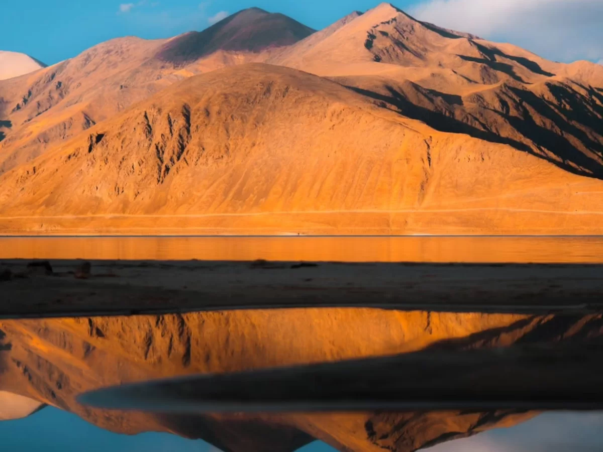 Ladakh Pangong Tso Lake sunset golden orange light terracotta brown rugged Himalayan mountains perfect reflection in calm turquoise blue brackish water clear sky high altitude scenic landscape adventure tour package. 