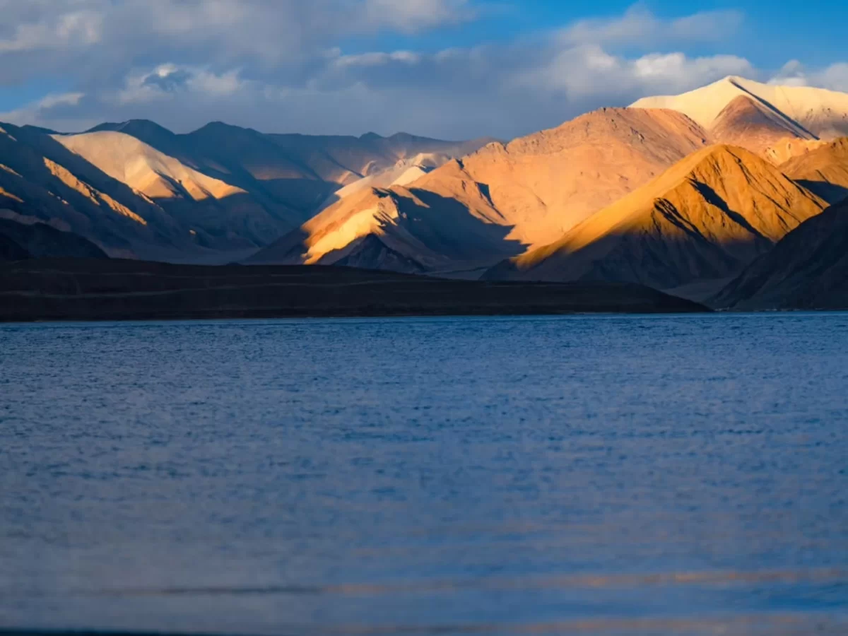 Ladakh Pangong Tso Lake golden hour soft sunlight on rugged brown Himalayan mountains blue brackish water foreground dramatic clouds high altitude scenic landscape adventure tour package Ladakh Himalayas.
