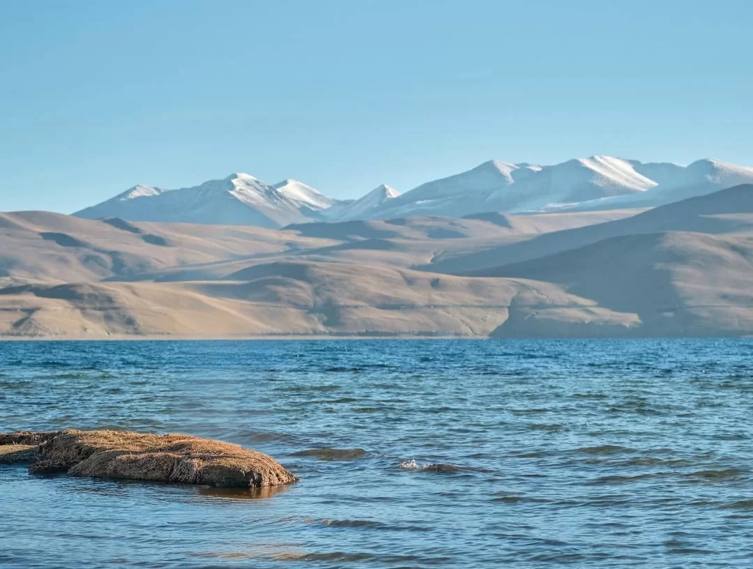 Ladakh Pangong Tso Lake deep blue brackish water snow capped jagged Himalayan mountains arid brown rocky barren slopes sandy shore clear pale blue sky high altitude adventure tour package Ladakh Himalayas.