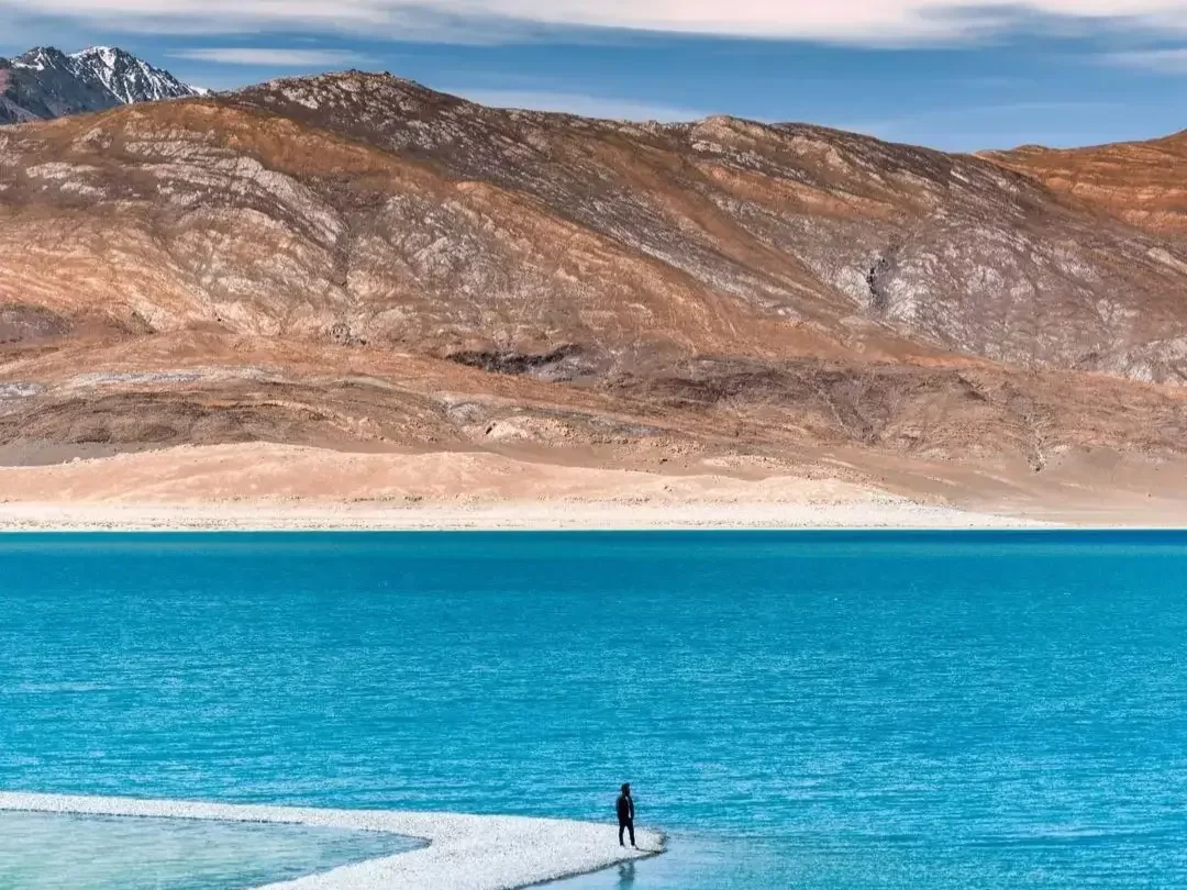 Ladakh Pangong Tso Lake turquoise blue high altitude lake snow peaked Himalayan mountains terracotta brown rocky arid slopes solo black silhouetted person white salt flat shoreline blue sky adventure tour package.