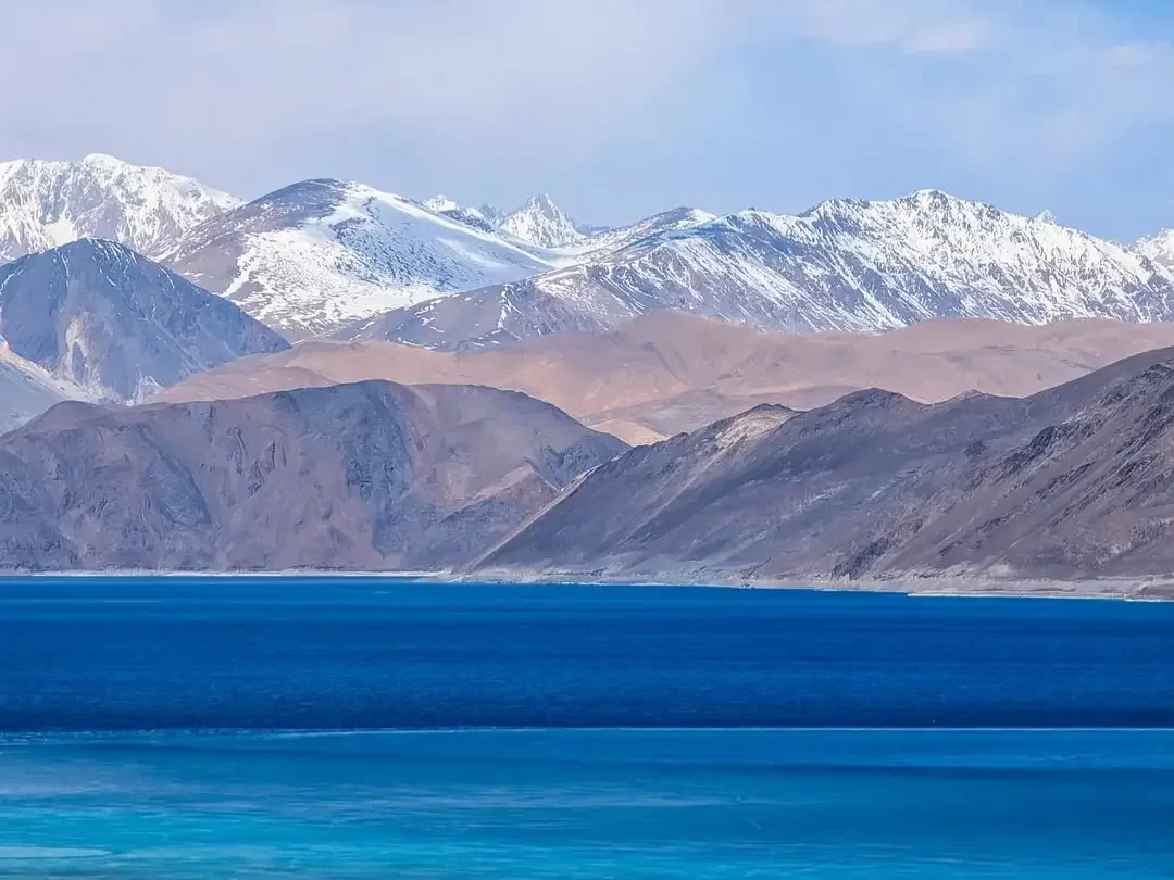 Ladakh Pangong Tso Lake deep blue turquoise brackish water snow capped rugged Himalayan mountains terracotta brown barren slopes scattered clouds pale blue sky high altitude scenic landscape adventure tour package