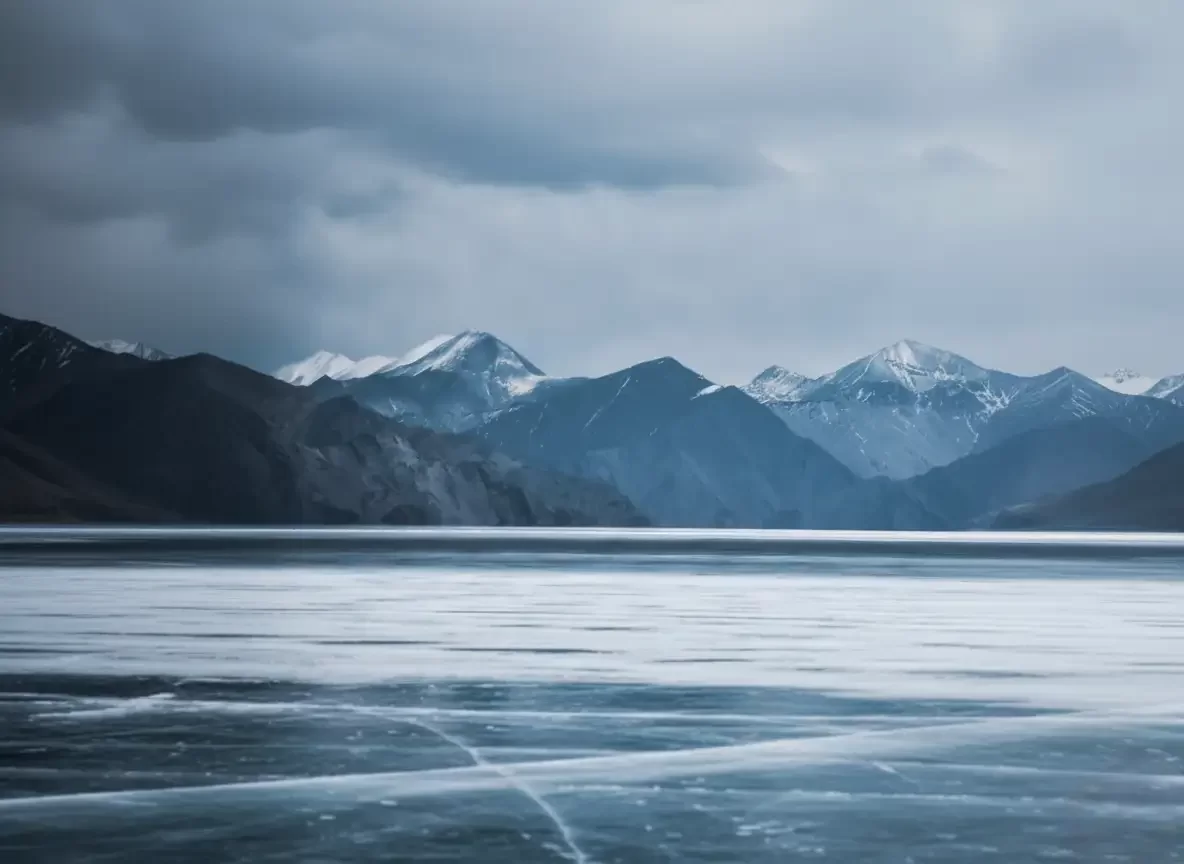 Pangong Lake at Pangong Tso Ladakh during overcast winter, featuring frozen surface cracks and distant snow mountains, perfect adventure Ladakh tour package. 