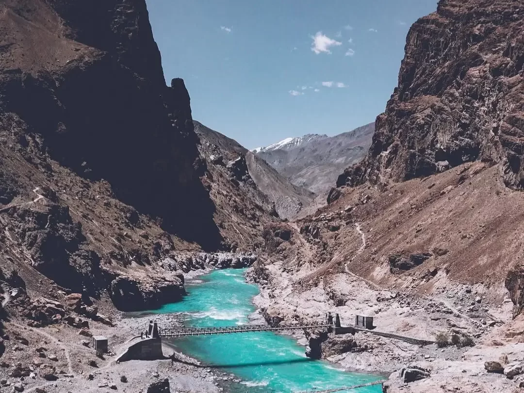 Turquoise Tsarap River flowing through rocky Zanskar gorge with steel suspension Purney Bridge near Purne village Ladakh India flanked by barren brown cliffs distant snowy peaks, perfect Spiti Zanskar tour package.