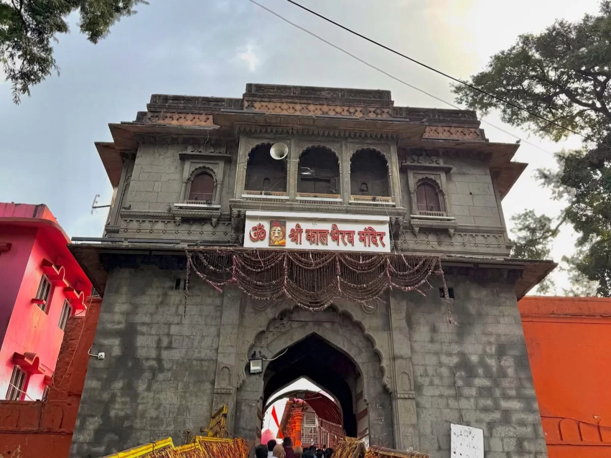 Shree Kaal Bhairav Temple gate at Ujjain during clear daylight, featuring grey stone arch red signboard garlands, perfect spiritual Madhya Pradesh tour package.