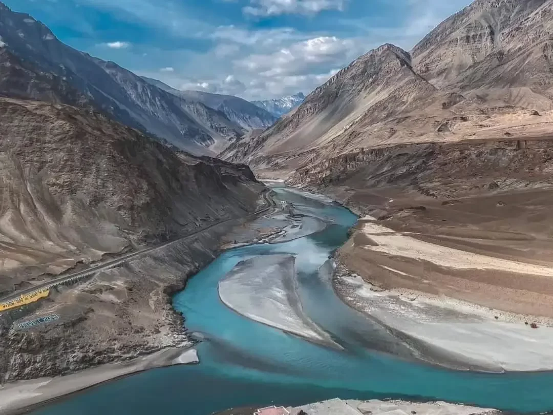 Sangam river confluence at Leh Ladakh during partly cloudy day, featuring turquoise waters valley mountains road bends, perfect adventure Ladakh tour package.
