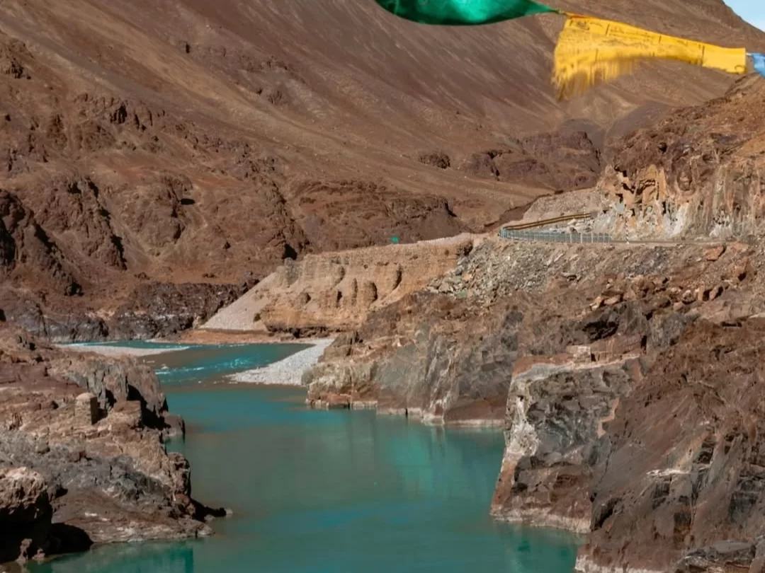 Prayer flags at Sangam Leh Ladakh during golden hour, featuring turquoise river gorge mountains road yellow rocks, perfect adventure Ladakh tour package.