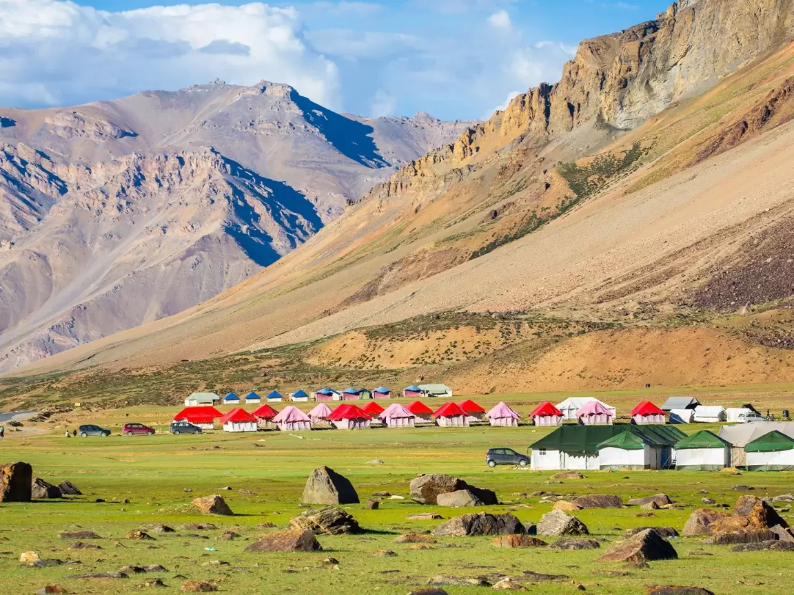 Colorful tent camps at Sarchu on Manali-Leh Highway during partly cloudy day, featuring green meadows, rugged mountains, perfect Ladakh tour package.