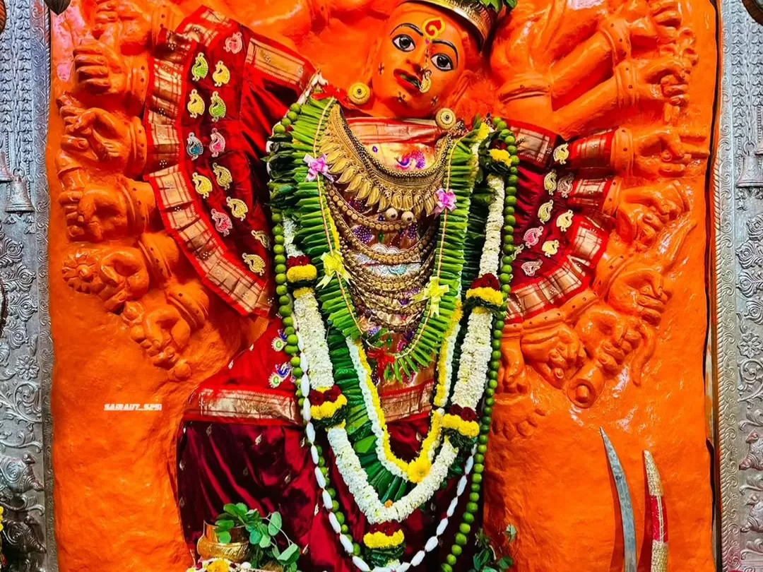 Shree Saptashrungi Nivasini Devi Shaktipeeth idol in Nashik adorned with red saree gold jewelry and floral garlands against vibrant orange carved backdrop