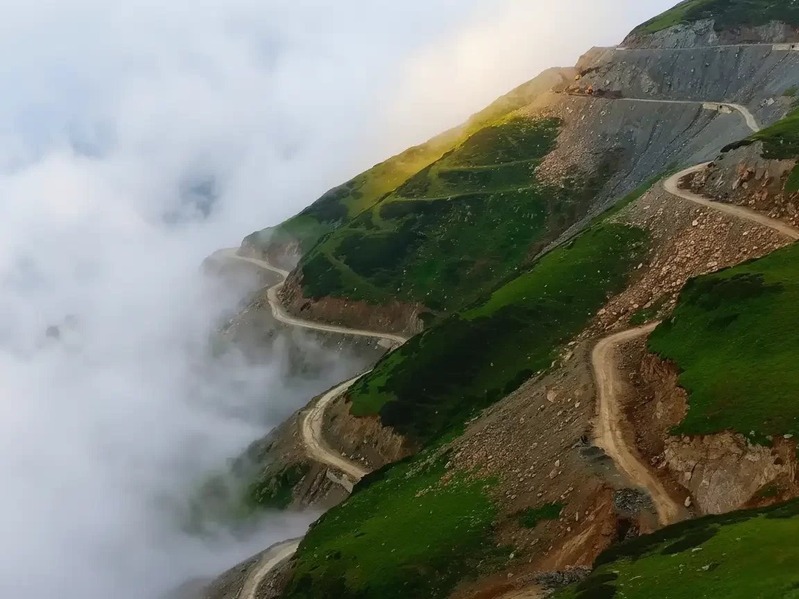 Sinthan Top misty mountain passes with winding roads & sun rays piercing clouds over green slopes, perfect Kashmir adventure tour packages.