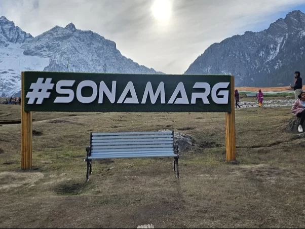 #Sonamarg sign at meadows during sunny day, featuring snowy mountains & bench, perfect adventure experience Kashmir tour packages.
