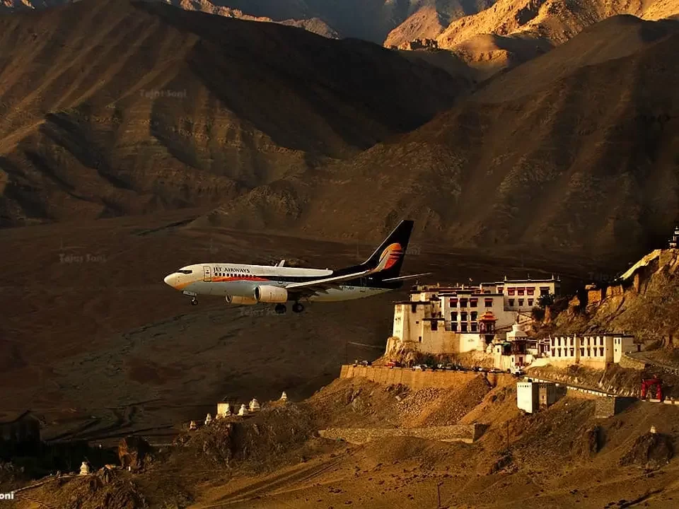 Spituk Monastery Ladakh dramatic low-altitude IndiGo jet landing over historic whitewashed gompa complex perched on rocky hilltop golden hour sunlight rugged brown mountains, breathtaking aviation Himalayan spiritual heritage tour package.