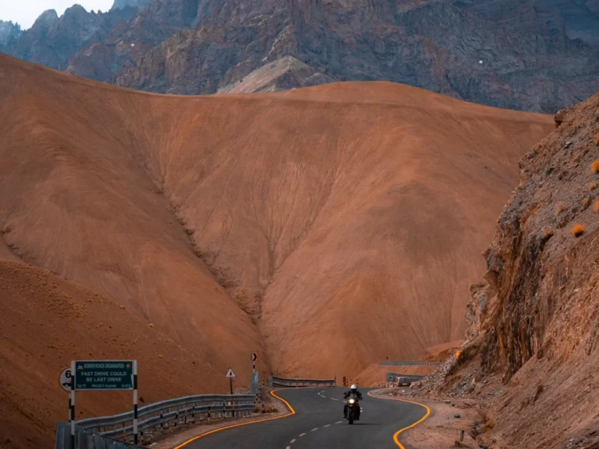Motorcycle rider on curving Srinagar Leh Highway through red sand dunes canyon near Mulbekh Ladakh during cloudy day, featuring highway sign towering rugged mountains, perfect biking adventure experience with Srinagar Leh Highway tour package. 