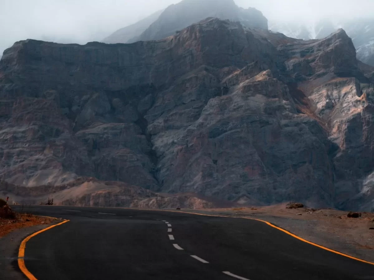 Curving Srinagar Leh Highway asphalt road through dramatic rocky canyon near Lamayuru Ladakh during foggy overcast day, featuring towering gray mountains distant peaks travelers, perfect adventurous drive experience with Srinagar Leh Highway tour package.
