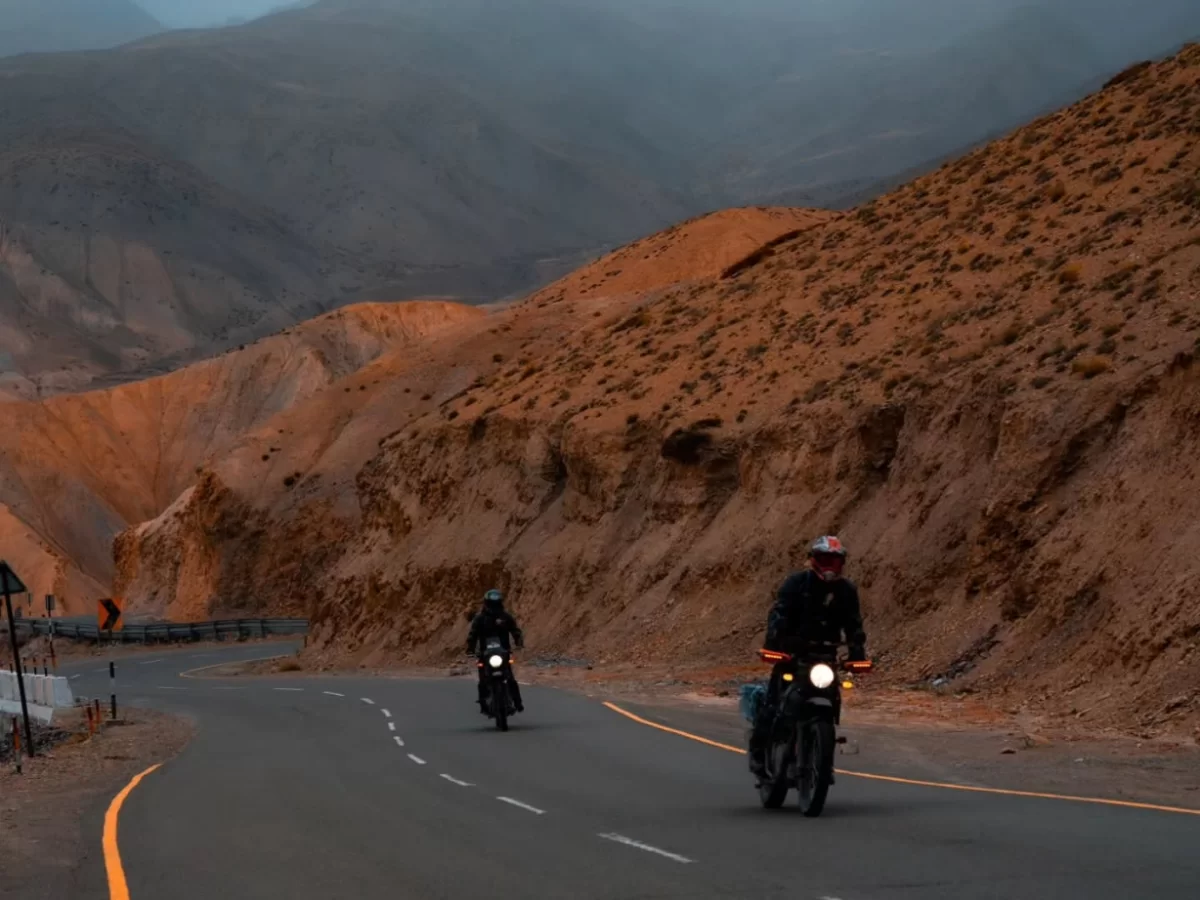 Two motorbikes riding on curving Srinagar Leh Highway through red rock canyon near Lamayuru Ladakh during twilight dusk, featuring rugged terracotta hills distant peaks road barriers, perfect biking adventure experience with Srinagar Leh Highway tour pack