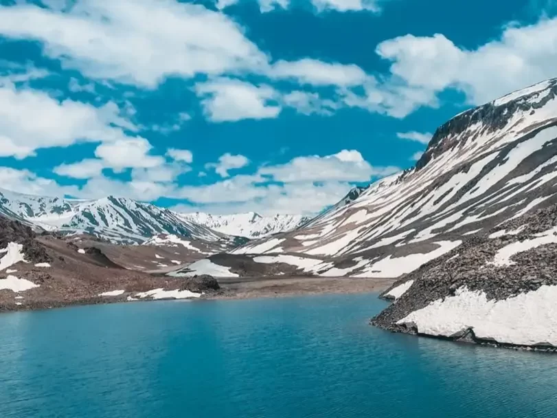 Suraj Tal Lahaul Spiti Himachal Pradesh India vibrant turquoise glacial lake amid snow capped peaks brown rocky mountains scattered white patches blue skies fluffy clouds foreground barren terrain, perfect Manali Leh highway high altitude tour package