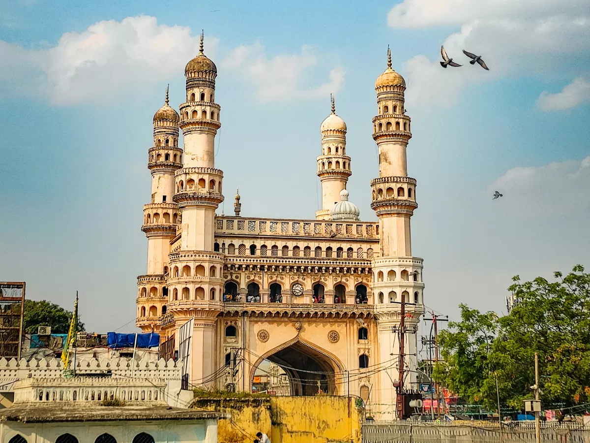 Front view of Charminar at Hyderabad featuring four minarets intricate arches birds flying under partly cloudy skies, ideal for Telangana tour packages.