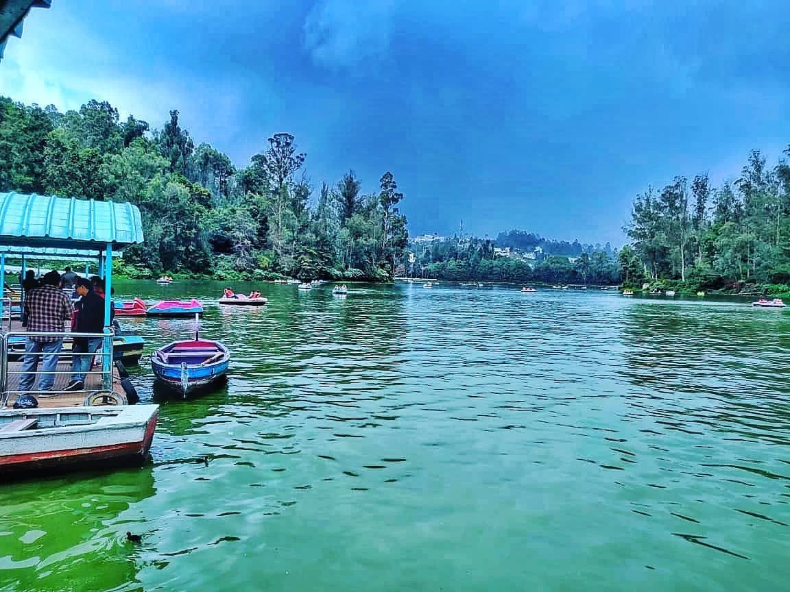 Busy Ooty Lake Udhagamandalam Nilgiris Tamil Nadu India jetty scene with tourists under blue tin roof shelter colorful pedal boats docked rowboats on emerald waters eucalyptus shola forests distant hills partly cloudy skies, perfect Nilgiri hill station b