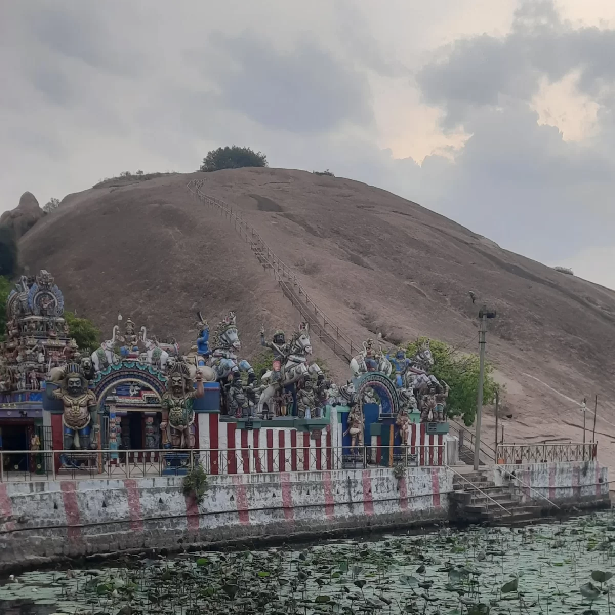 Samanar Hill Tamil Nadu Samanamalai Jain temple base colorful gopuram towers deities horses steps brown rocky hill lotus pond cloudy sky lamp post railing, ancient Jain temple hill temple tour package. 