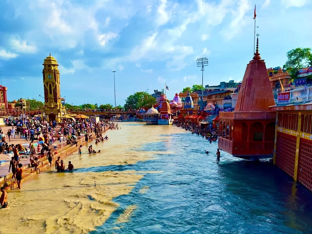 Har Ki Pauri Haridwar vibrant Ganga ghats with golden clock tower temples devotees bathing colorful umbrellas blue sky clouds, perfect spiritual bathing ghat, Uttarakhand tour packages.
