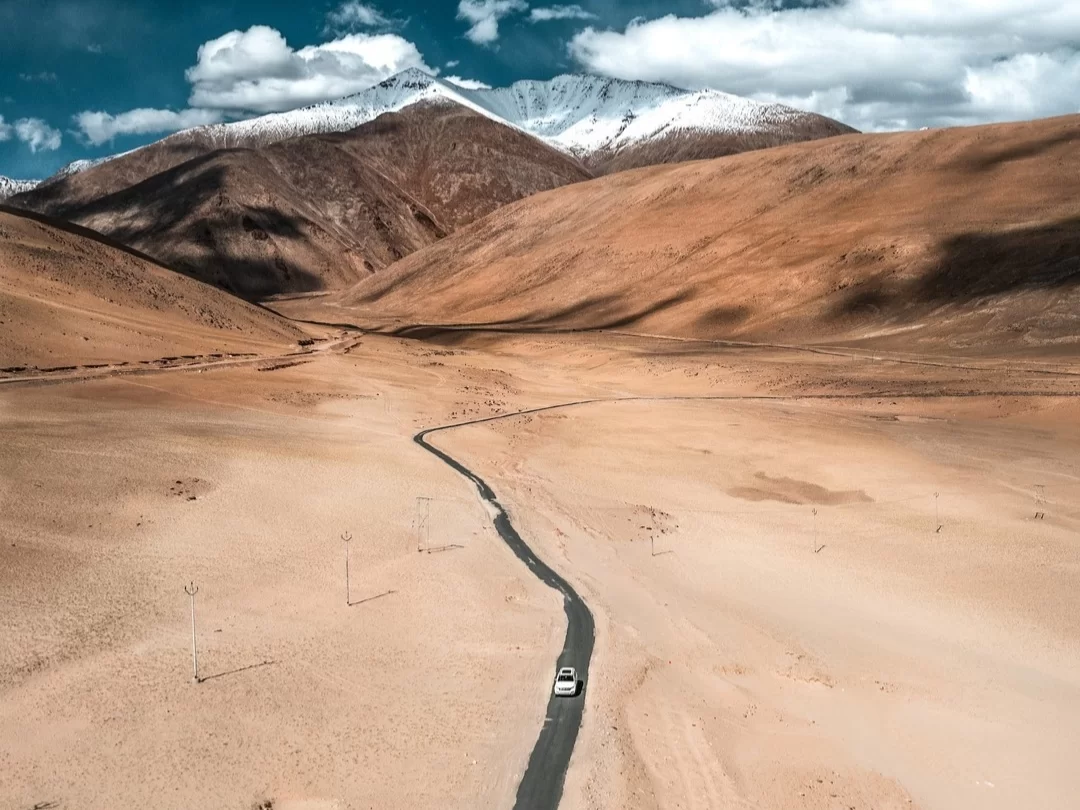 Drone aerial view of lone white car driving snaking black road through vast golden sandy desert valley flanked by terracotta brown barren hills snow-capped peaks under partly cloudy blue sky near Tso Kar Ladakh India, perfect high-altitude road trip adven