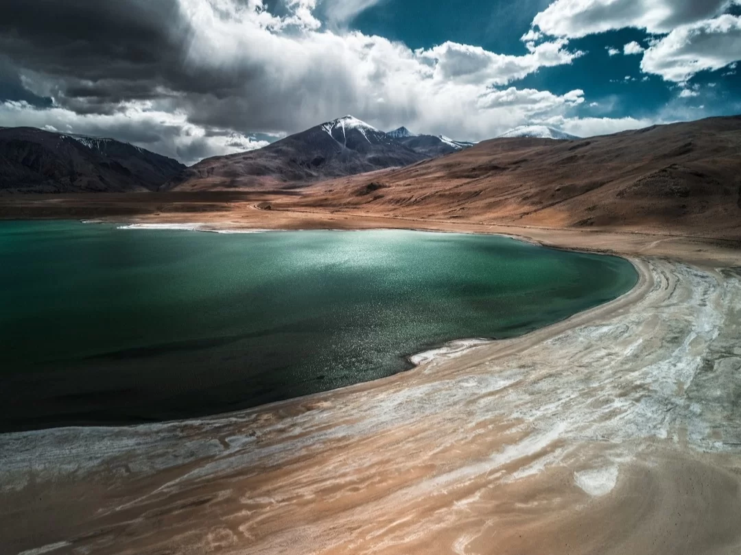 Aerial view of vibrant turquoise crescent Tso Kar lake with white sandy shores terracotta brown hills snow-capped peaks dramatic stormy cloudy sky Ladakh India, perfect Ramsar wetland scenic photography adventure with Leh Ladakh tour package.