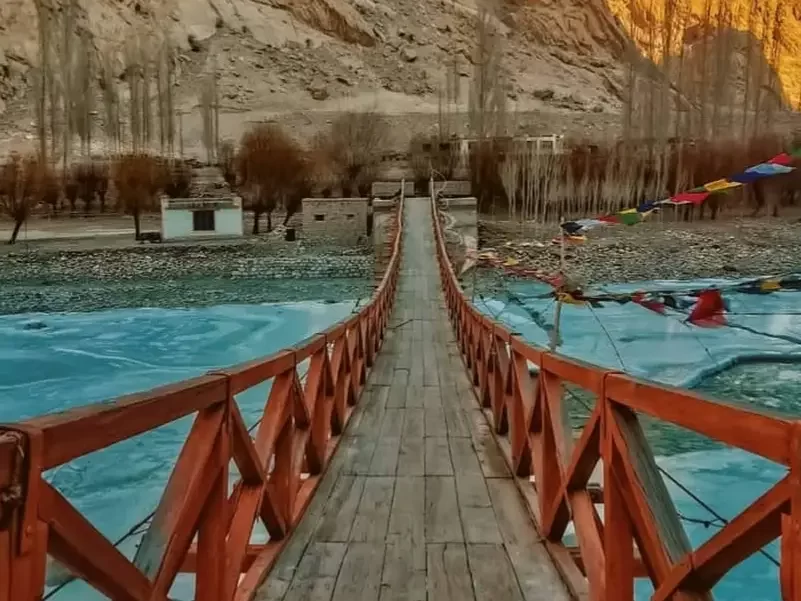 Red wooden hanging bridge over turquoise river near Turtuk village Nubra Valley Ladakh during golden hour sunset, featuring prayer flags, barren mountains, perfect adventure Turtuk tour package