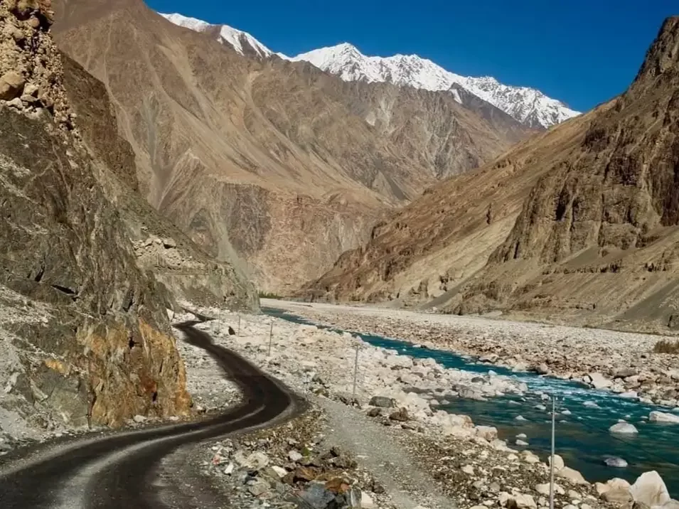 Scenic winding road along Shyok River to Turtuk village Nubra Valley Ladakh during clear blue sky, featuring snow-capped Karakoram peaks, barren gorges, perfect adventure Turtuk tour package. 