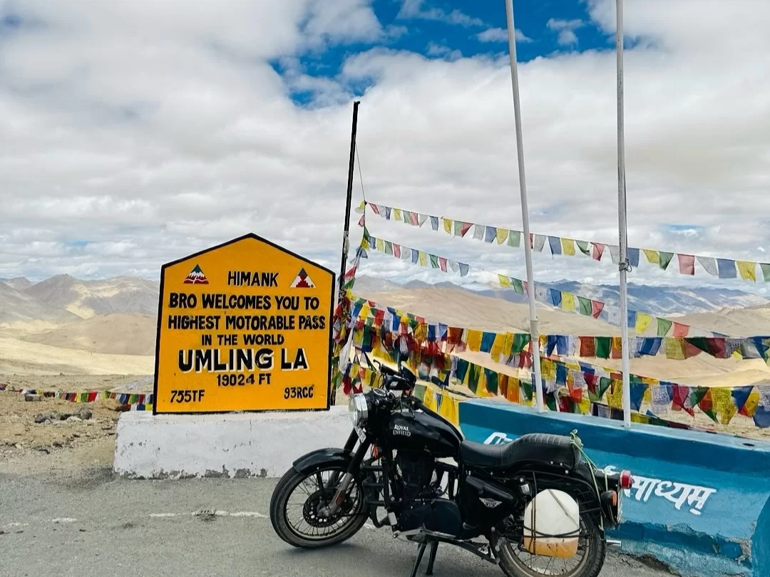 Motorcycle at Umling La Pass Ladakh world's highest motorable pass under cloudy skies, featuring prayer flags and mountains, perfect adventure Ladakh tour package. ​