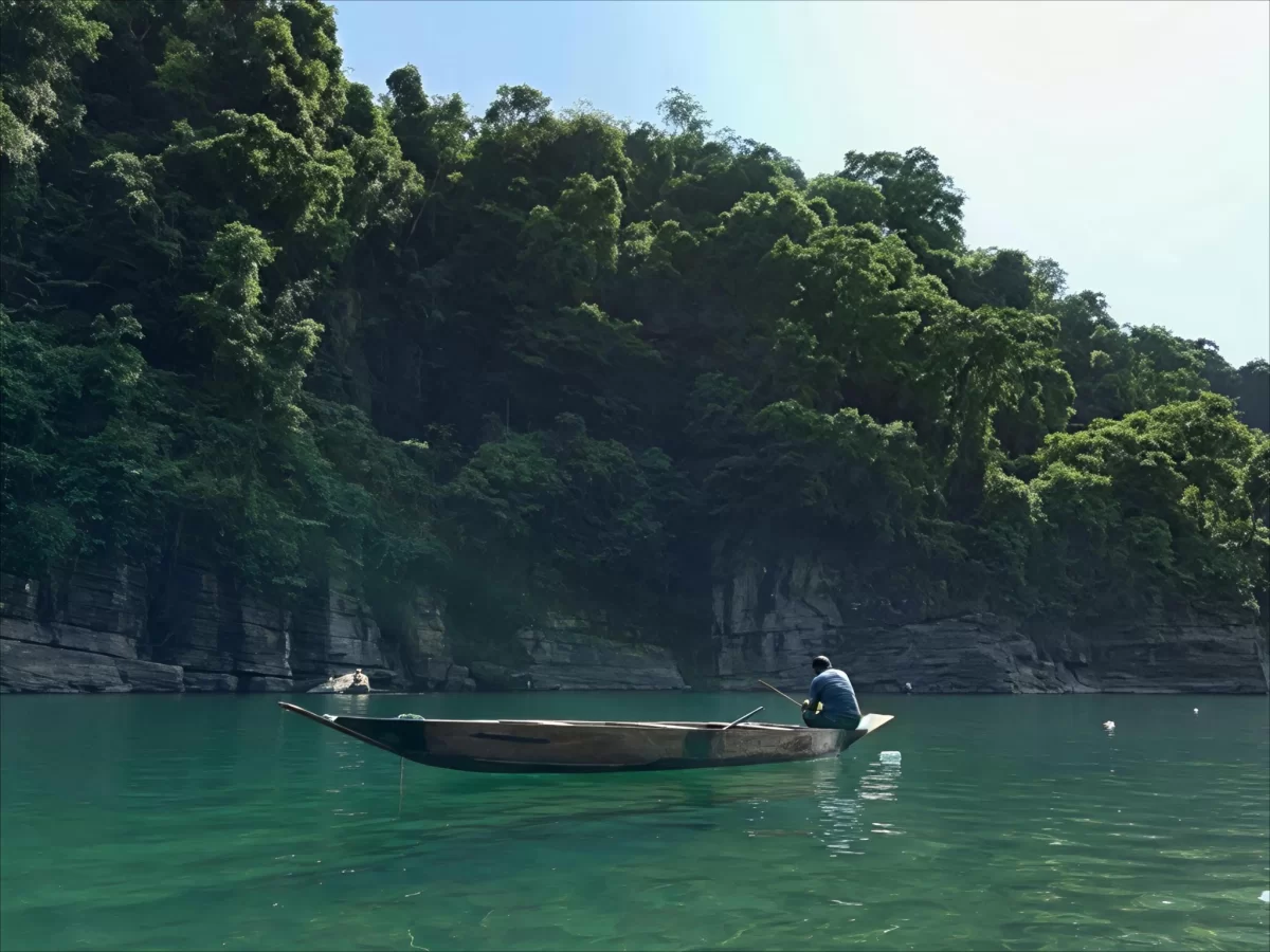 Umngot River Dawki Meghalaya fisherman traditional wooden boat lush green forested cliffs rocky banks crystal clear emerald waters sunny day serene tranquil vibe perfect boating fishing adventure Meghalaya tour package.